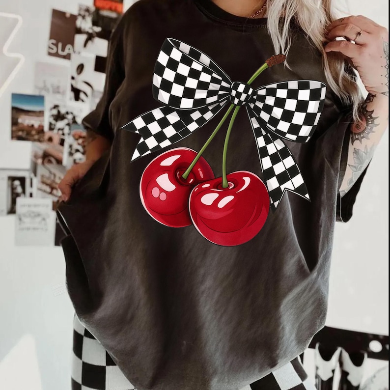 Puede incluir: Camiseta gris oscuro con un gr&aacute;fico de dos cerezas rojas brillantes, tallos verdes y un lazo a cuadros blanco y negro. La camiseta tiene un corte holgado y mangas cortas.