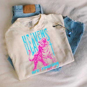 May include: Cream-colored t-shirt with a vibrant graphic of a roaring pink tiger and the words "HEAVENS ARE ROARING" in turquoise. The text "PSALM 19:1" is also visible. The shirt is paired with light blue denim jeans and round-framed glasses.
