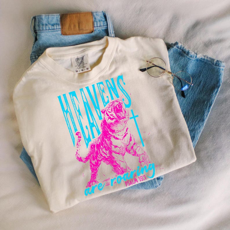 May include: Cream-colored t-shirt with a vibrant graphic of a roaring pink tiger and the words "HEAVENS ARE ROARING" in turquoise. The text "PSALM 19:1" is also visible. The shirt is paired with light blue denim jeans and round-framed glasses.