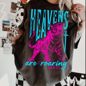 May include: Dark gray t-shirt featuring a vibrant pink tiger graphic with the words "HEAVENS" in blue above it. Below the tiger, the text "are roaring" is displayed in blue, with "PSALM 19:1" underneath.