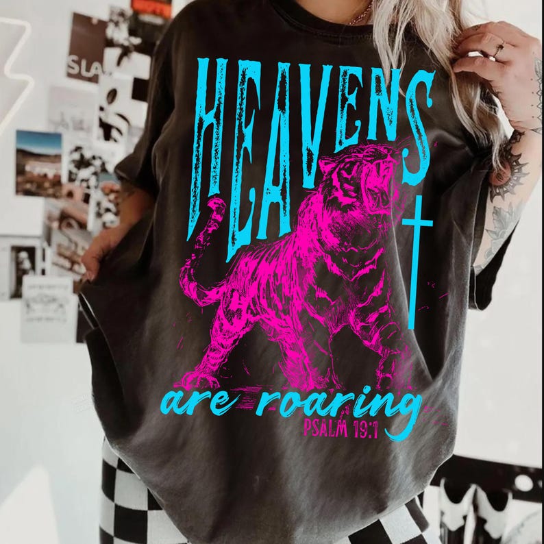 May include: Dark gray t-shirt featuring a vibrant pink tiger graphic with the words "HEAVENS" in blue above it. Below the tiger, the text "are roaring" is displayed in blue, with "PSALM 19:1" underneath.