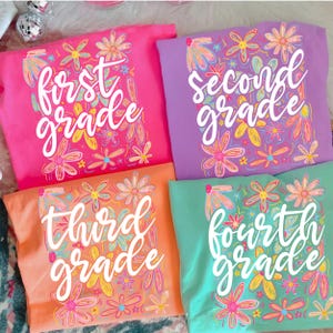 May include: Four colorful t-shirts, each displaying a grade level: First, Second, Third, and Fourth. The shirts are pink, purple, orange, and teal, adorned with floral designs in various colors. The grade level is written in white script.