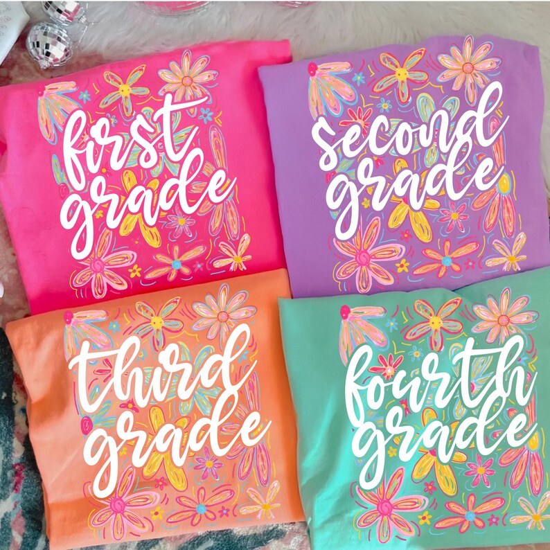 May include: Four colorful t-shirts, each displaying a grade level: First, Second, Third, and Fourth. The shirts are pink, purple, orange, and teal, adorned with floral designs in various colors. The grade level is written in white script.