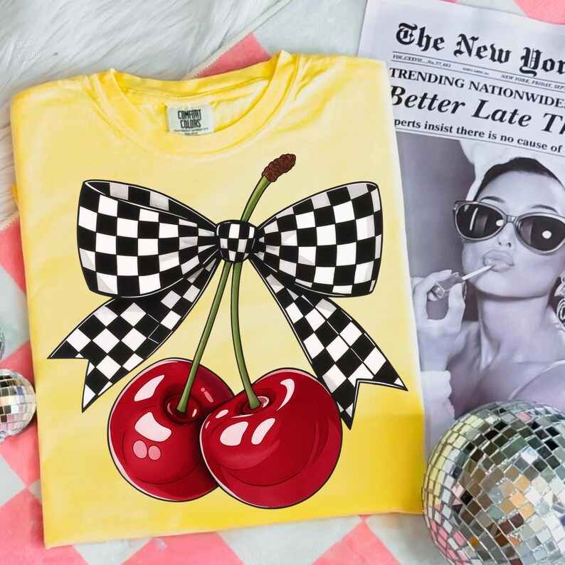 Puede incluir: Camiseta amarilla Comfort Colors con un gr&aacute;fico de dos cerezas rojas y un lazo a cuadros blanco y negro. Se ve un peri&oacute;dico con el titular "TRENDING NATIONWIDE Better Late T". Una bola de discoteca est&aacute; en el fondo.