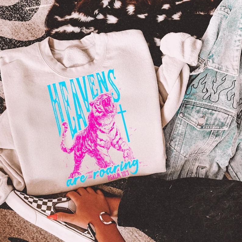 May include: Cream-colored sweatshirt with a vibrant pink tiger graphic and the text "HEAVENS are roaring PSALM 19:1" in blue. The sweatshirt is paired with a denim jacket and checkered slip-on shoes.