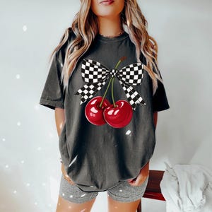 Puede incluir: Camiseta gris oscuro con un gr&aacute;fico de dos cerezas y un lazo a cuadros. Las cerezas son de un rojo vibrante con tallos verdes. El lazo es a cuadros blanco y negro. La camiseta es de estilo casual.