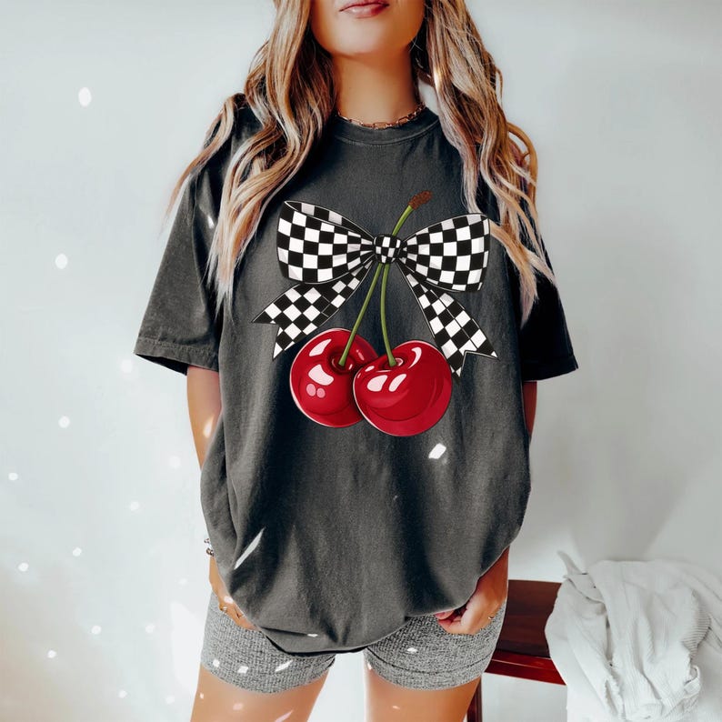 Puede incluir: Camiseta gris oscuro con un gr&aacute;fico de dos cerezas y un lazo a cuadros. Las cerezas son de un rojo vibrante con tallos verdes. El lazo es a cuadros blanco y negro. La camiseta es de estilo casual.