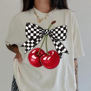 Puede incluir: Camiseta color crema con un gr&aacute;fico de dos cerezas rojas brillantes con tallos verdes, atadas con un lazo a cuadros blanco y negro. El dise&ntilde;o est&aacute; centrado en la parte delantera, creando un aspecto l&uacute;dico y llamativo.