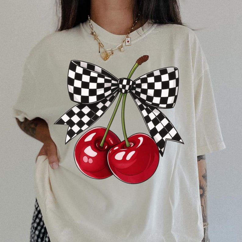 Puede incluir: Camiseta color crema con un gr&aacute;fico de dos cerezas rojas brillantes con tallos verdes, atadas con un lazo a cuadros blanco y negro. El dise&ntilde;o est&aacute; centrado en la parte delantera, creando un aspecto l&uacute;dico y llamativo.