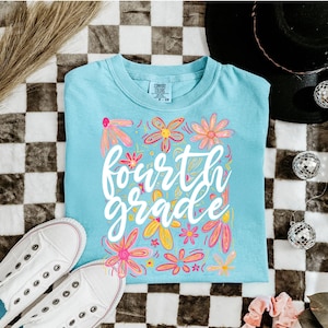 May include: A light blue t-shirt with the words "fourth grade" in white script, surrounded by colorful floral designs. The shirt is paired with white sneakers on a checkered background.
