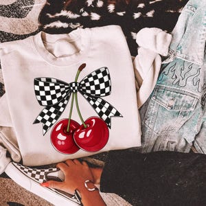 Puede incluir: Sudadera color crema con un gr&aacute;fico de dos cerezas rojas brillantes con tallos verdes y un lazo a cuadros blanco y negro. Parte de una colecci&oacute;n de ropa casual.