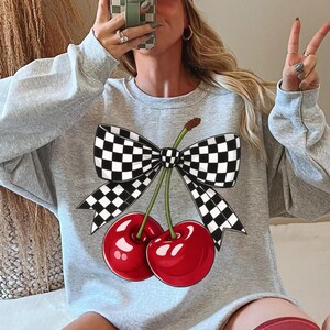 Puede incluir: Una sudadera gris claro con un gr&aacute;fico de dos cerezas rojas brillantes y un lazo a cuadros blanco y negro. La sudadera tiene cuello redondo y mangas largas. El dise&ntilde;o est&aacute; centrado en la parte delantera.