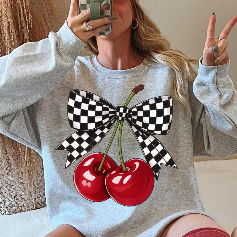 Puede incluir: Una sudadera gris claro con un gr&aacute;fico de dos cerezas rojas brillantes y un lazo a cuadros blanco y negro. La sudadera tiene cuello redondo y mangas largas. El dise&ntilde;o est&aacute; centrado en la parte delantera.
