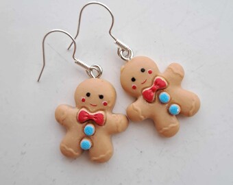 May include: A pair of gingerbread man earrings. Each earring features a tan gingerbread man with a red bow tie, blue buttons, and a smiling face. The earrings have silver-coloured fish hook ear wires.