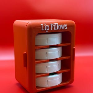 May include: An orange storage unit with the text Lip Pillows printed in white on the top front. The unit features four horizontal shelves, each holding a round white container. The structure has a matte finish and a rectangular shape.