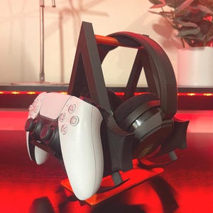 Puede incluir: Un soporte de impresión 3D negro con un auricular y un mando de videojuego blanco. El soporte tiene una forma triangular y un asa negra y naranja.