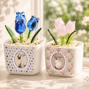 May include: Two square white ceramic planters filled with small stones and glass tulips. The left planter features blue tulips and a label with the name Bella and a cat image. The right planter has pink tulips and a label with the name Freya and a cat.