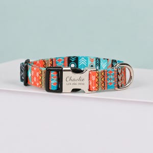 May include: A pet collar featuring a vibrant orange, teal, and black geometric pattern with arrows and diamonds. It includes a silver-toned metal buckle engraved with the name Chavlie and the phone number 123-456-7890, plus a black plastic adjuster.