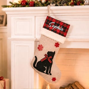 May include: A hessian Christmas stocking featuring a black cat silhouette with a red and black tartan bow. The cuff is white faux fur topped with a red and black tartan fabric embroidered with the name Sophia in white script and red snowflakes.