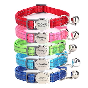 Może przedstawiać: Pięć kolorowych obroży dla kotów z srebrnymi dzwoneczkami i spersonalizowanymi etykietami z imionami. Obroże są czerwone, różowe, zielone, niebieskie i ciemnoniebieskie. Etykiety z imionami zawierają napisy "Cookie", "Ginger", "Happy", "Simba" i "Whiskers" wraz z numerami telefonów.