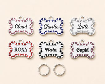 May include: Six bone-shaped metal pet ID tags with a loop at the top. Each tag features a border of colored rhinestones and a custom name engraved in black text: Cloud, Charlie, Latte, ROXY, Rain, and Cupid. Two silver metal split rings are included.