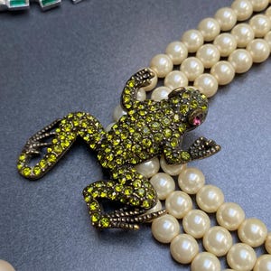 Op de afbeelding: Een vintage-stijl ketting met drie strengen imitatieparels. Een gedetailleerde kikkerhanger, versierd met groene strass en een roze oog, ligt op de parels. Andere sieraden zijn zichtbaar op de achtergrond.