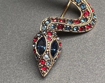 May include: A gold-toned snake brooch with a detailed design. The snake's body is adorned with red and blue rhinestones, and the head features two large blue stones for eyes. The brooch is a decorative accessory.