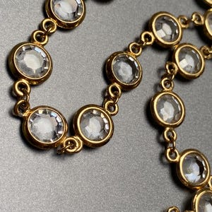 May include: Close-up of a gold necklace with clear, round gemstones. The gemstones are set in individual bezels and linked with small gold connectors. The necklace is shown against a grey background.