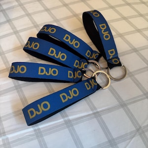 以下が含まれることがあります： 金色の「DJO」の文字が入った青いキーホルダー。キーホルダーは黒い裏地とシルバーのキーリングが付いています。いくつかのキーホルダーが白とグレーのチェック柄の表面に配置されています。