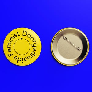 Op de afbeelding: Een gele knop met een zwarte cirkel en een pijl in het midden. De tekst rond de cirkel luidt "Doorgetraaide Feminist". De achterkant van de knop is zilverkleurig met een pin.