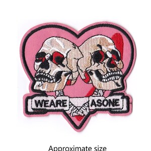 May include: Pink embroidered patch with two skulls facing each other inside a heart. The skulls have red eyes and are holding a banner that says "We Are As One". The patch is approximately 3.5 inches by 3.25 inches.