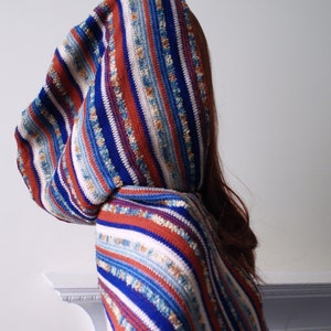 May include: A hooded poncho with a striped pattern in shades of blue, red, white and beige. The poncho is made of a soft, textured fabric.