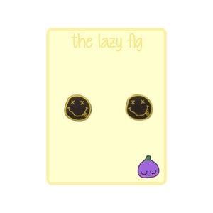 Pode incluir: Um par de brincos de cara sorridente dourados e pretos com um 'X' para os olhos e uma l&iacute;ngua para fora. Os brincos est&atilde;o sobre um fundo amarelo com o texto 'the lazy fig' no topo e uma figueira roxa de desenho animado com uma cara sorridente na parte inferior.