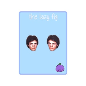 May include: A pair of earrings featuring a photo of a man with brown hair. The earrings are on a light blue background with the text "the lazy fig" at the top and a cartoon purple fig with a smiling face at the bottom.