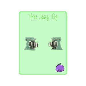 May include: A pair of pastel green stand mixer earrings with a silver bowl and a purple fig with a smiling face on a green background with the text "the lazy fig"