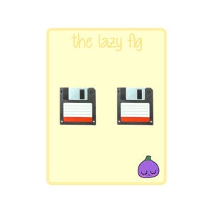 Puede incluir: Dos pendientes de disquete negros y rojos sobre un fondo amarillo claro. El texto "the lazy fig" está en la parte superior. Una higo morado con una cara sonriente está en la parte inferior derecha.