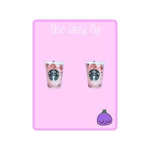 May include: A pair of pink and white Starbucks Frappuccino earrings with the Starbucks logo on the front. The earrings are on a pink background with the text "the lazy fig" at the top and a cartoon purple fig with a smiling face at the bottom.