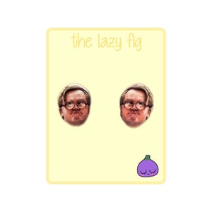 Puede incluir: Un par de pendientes con una imagen de primer plano de un hombre con gafas y una expresión seria. Los pendientes son redondos y tienen un fondo blanco. El texto "the lazy fig" está impreso sobre un fondo amarillo por encima de los pendientes. Una higuera de dibujos animados morada con una cara sonriente está impresa debajo de los pendientes.