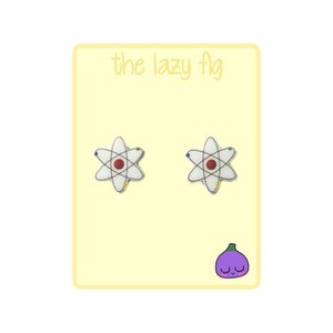 May include: A pair of white atom earrings with a red center and blue and yellow orbiting electrons. The earrings are on a yellow background with the text "the lazy fig" and a purple fig with a smiling face.