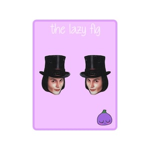 May include: A pair of earrings featuring a cartoon image of Willy Wonka from the movie "Willy Wonka and the Chocolate Factory". The earrings are on a pink background with the text "the lazy fig" at the top and a cartoon purple fig with a smiling face at the bottom.