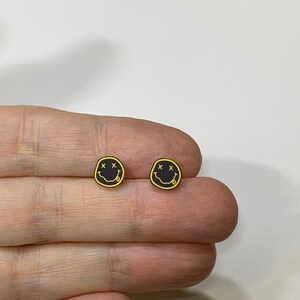 Pode incluir: Um par de brincos de bot&atilde;o com carinhas sorridentes pretas e amarelas. As carinhas sorridentes t&ecirc;m um 'X' para os olhos e uma l&iacute;ngua de fora.