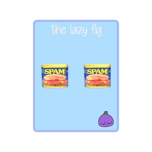 May include: A pair of earrings featuring a miniature Spam can with a sandwich inside. The earrings are on a light blue background with the text "the lazy fig" at the top and a purple fig with a face at the bottom.
