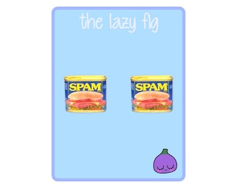 Può includere: Un paio di orecchini con una lattina di Spam in miniatura con un panino al suo interno. Gli orecchini sono su uno sfondo blu chiaro con il testo "the lazy fig" in alto e un fico viola con una faccia in basso.