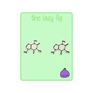 Op de afbeelding: Een paar witte oorbellen met een geprinte chemische structuur van cafeïne. De oorbellen zijn op een groene achtergrond met de tekst "the lazy fig" bovenaan en een cartoon paarse vijg met een glimlachend gezicht onderaan.