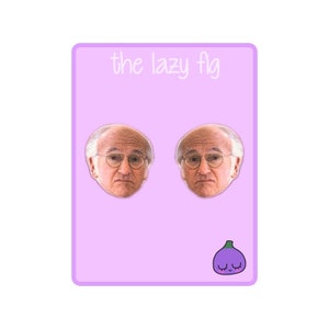 Op de afbeelding: Een paar oorbellen met een cartoonafbeelding van een mannengezicht. De oorbellen bevinden zich op een roze achtergrond met de tekst "the lazy fig" bovenaan. Een cartoonvijgenboom met een paarse body en een groene steel bevindt zich onderaan de afbeelding.