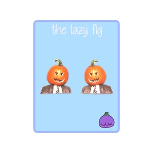 Puede incluir: Un par de pendientes con forma de calabaza con un traje negro y corbata. Los pendientes están sobre un fondo azul claro con el texto "the lazy fig" en la parte superior y una higo morado con una cara en la parte inferior.