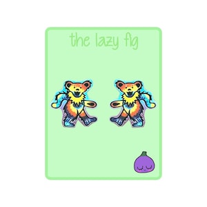 May include: A pair of tie-dye bear earrings with a purple, blue, orange, and yellow color scheme. The earrings are on a green background with the text "the lazy fig" and a purple fig with a face.