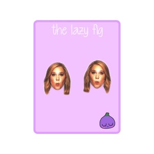 May include: A pair of earrings featuring a close-up image of a woman's face with a surprised expression. The earrings are on a pink background with the text "the lazy fig" at the top and a cartoon purple fig with a smiling face at the bottom.