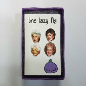 May include: A purple box with a white insert featuring four images of women with the text "the lazy fig" printed on the top. The bottom of the insert features a purple cartoon fig with a smiling face.
