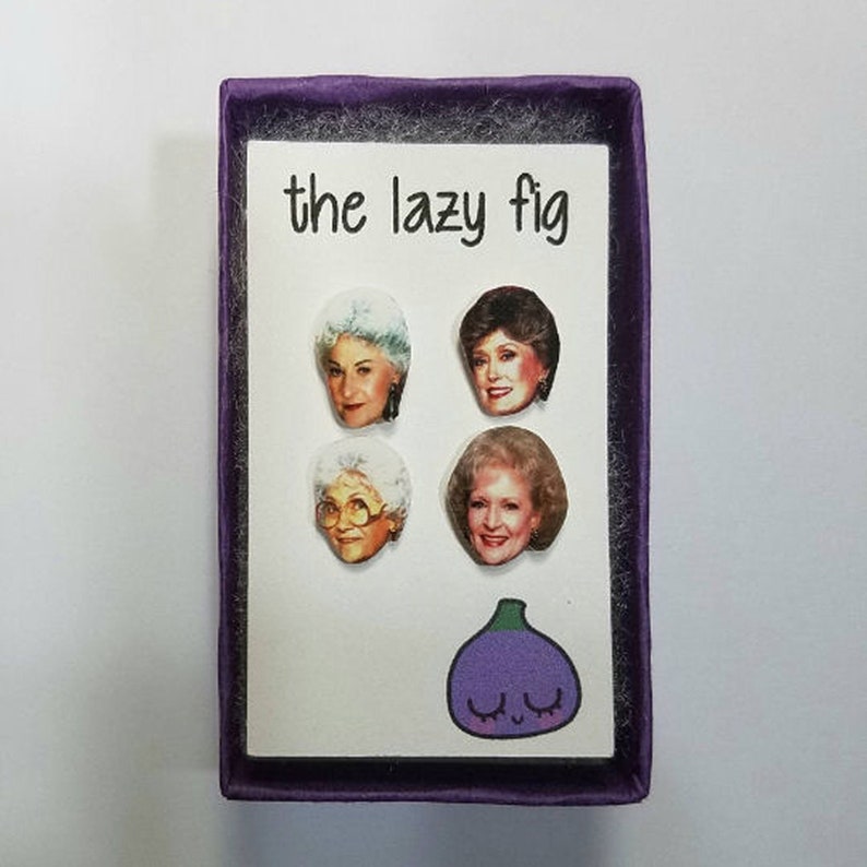 May include: A purple box with a white insert featuring four images of women with the text "the lazy fig" printed on the top. The bottom of the insert features a purple cartoon fig with a smiling face.